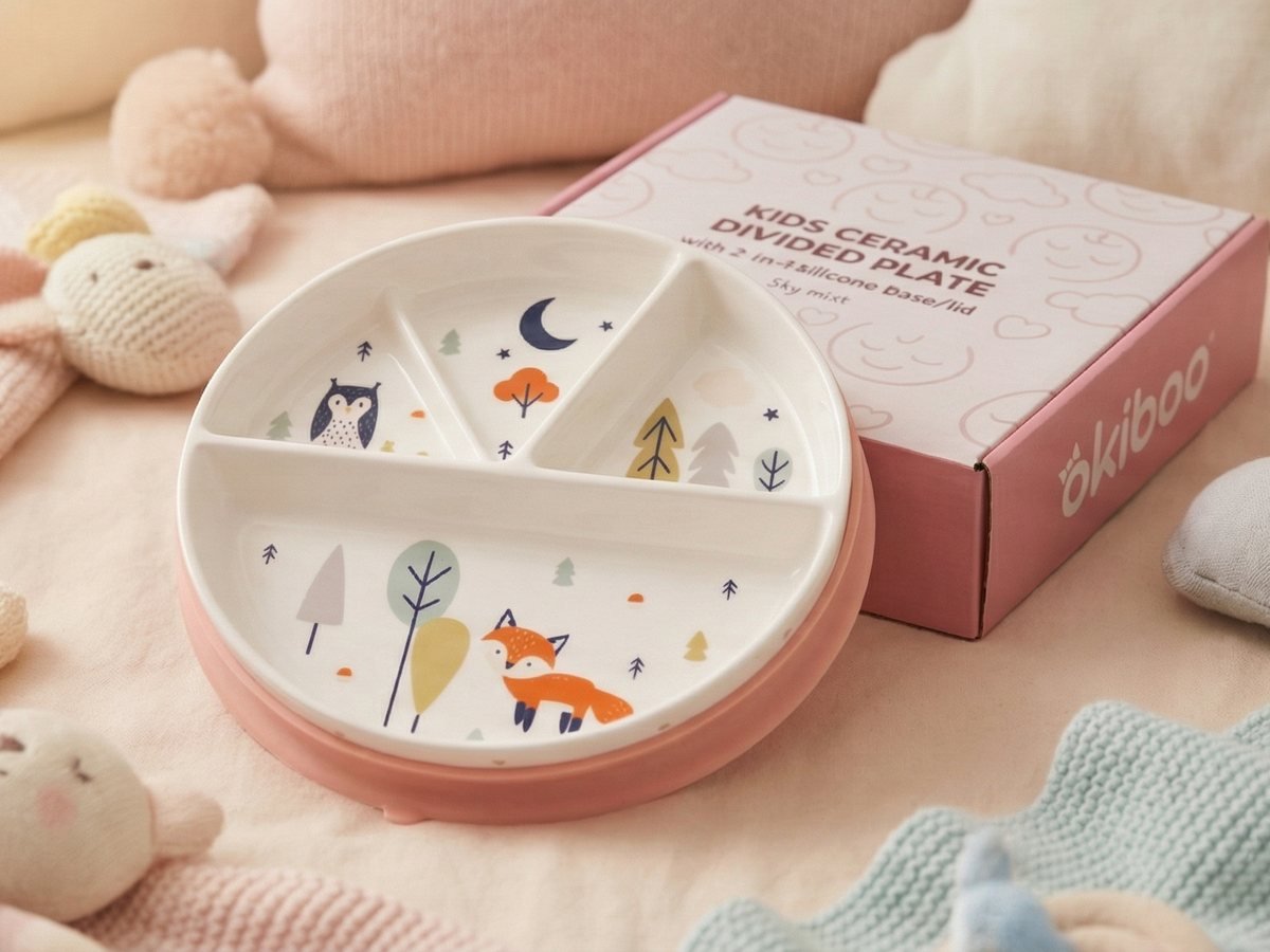 A Forest Friends plate in its peach silicone base beside its okiboo gift box, on a nursery blanket