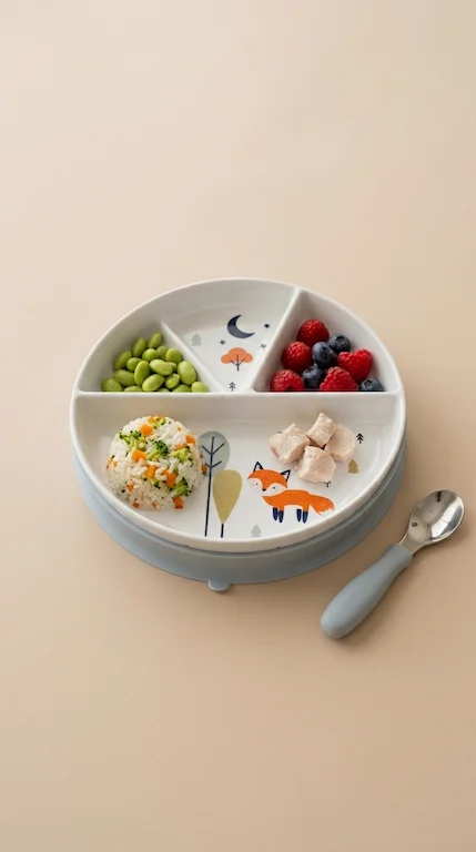 The plate sits on light oak with washed linen. Each section holds a small, separate portion that never touches the next: green peas, soft cubes of roasted sweet potato, a few blueberries, strips of toast.